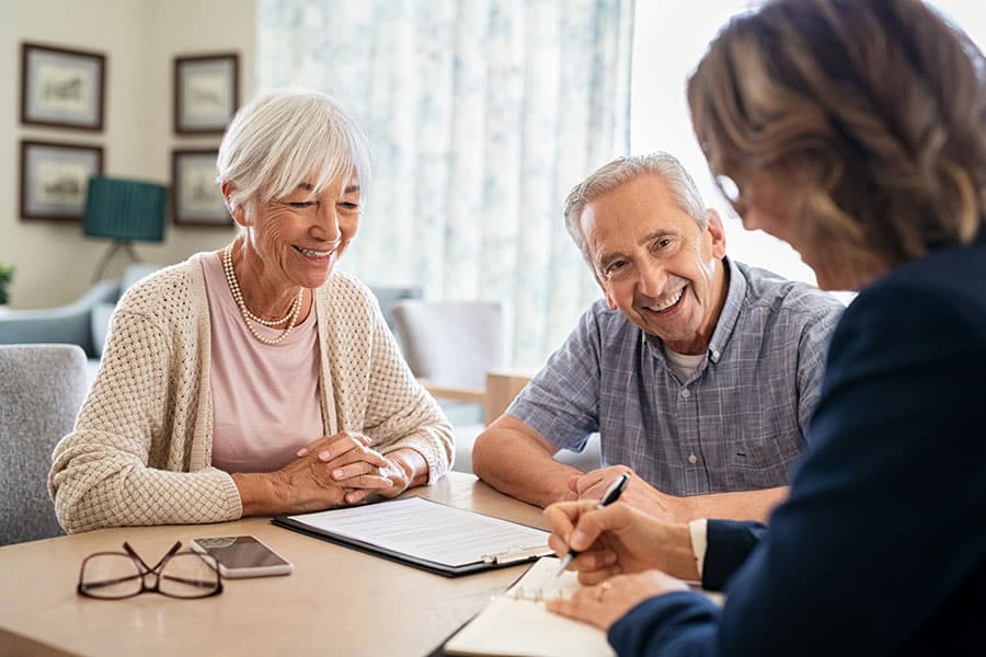 Smiling older couple sits at a table with a professional, reviewing documents together. The man is smiling at the camera while the woman listens attentively as they discuss options for bundling insurance. A clipboard and phone are on the table.