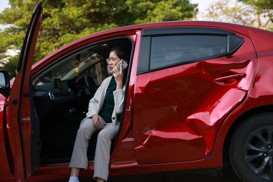 A woman sits in a red car with a large dent in the door, talking on her phone—perhaps discussing bundling insurance benefits after her recent mishap. The driver's door is open, and trees are visible in the background.