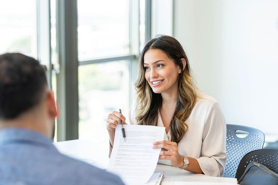 A woman sits at a table, smiling and holding paperwork while talking to another person across from her in a bright office setting, as they discuss the advantages of bundling insurance benefits.