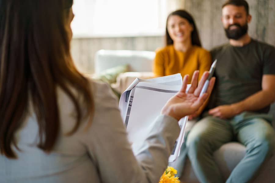 A woman holding documents speaks to a smiling couple sitting on a couch, suggesting a meeting or counseling session about protecting business in a comfortable, well-lit room.