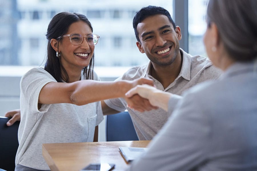 A smiling couple sits at a table, shaking hands with a person across from them in a professional setting—an ideal moment highlighting the importance of small business protection. The background shows large windows and a blurred cityscape.