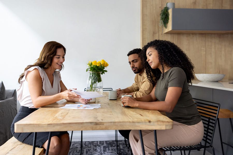 Three people sit around a wooden table in a modern kitchen, discussing documents related to small business policies. Two hold mugs, while one gestures to the papers outlining essential business protection strategies. A vase of yellow flowers sits on the table.