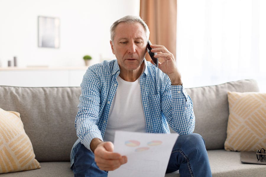 An older man sits on a sofa, talking on the phone while looking at a piece of paper with charts about insurance deductibles. He appears focused and concerned.