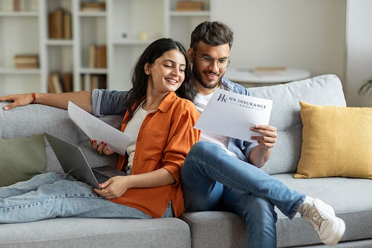 A smiling couple sits on a couch looking at insurance papers. The woman holds a laptop and leans on the man, who is holding a document labeled HN Insurance Group.