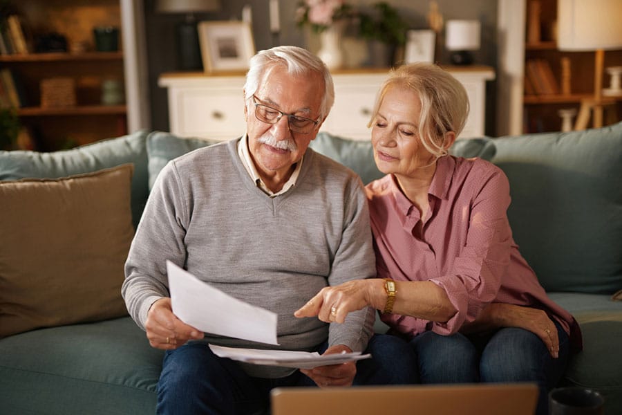 An elderly couple sits on a couch reviewing paperwork together, looking focused. The man holds papers while the woman points at them. A laptop is open in front of them on the table. The room appears cozy and warmly lit.