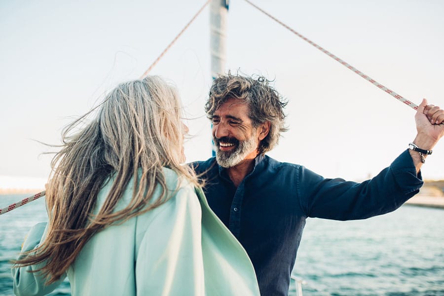 An older couple stands on a boat, smiling at each other. The man has curly gray hair and a beard, wearing a navy shirt, with arms stretched out; the woman has long gray hair and wears a light coat. The sea is in the background.