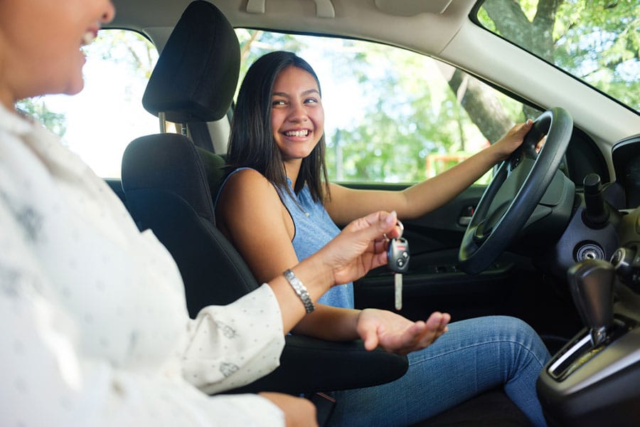 A smiling young woman sits in the driver’s seat of a car, reaching for car keys being handed to her by a person in the passenger seat. Sunlight streams through the windows, creating a cheerful atmosphere that perfectly captures an important moment for teen driver safety.