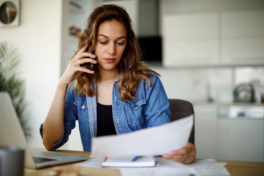 A woman in a denim shirt sits at a table with a laptop, papers, and a notebook, talking on her phone about insurance deductibles and looking at a document. The background shows a modern kitchen.
