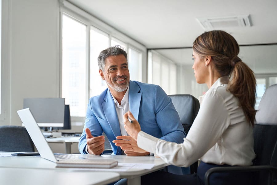 Two professionals sit at a desk in a modern Metro Detroit office, having a discussion. The man in a blue blazer smiles while the woman, holding a pen, speaks. A laptop and papers are on the desk—clear signs of dedicated insurance agents exploring tailored independent business insurance solutions.