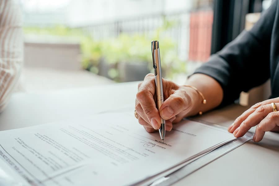 A person signs a document with a silver pen at a table—perhaps finalizing paperwork with Insurance Agents. Only their hand and part of their arm are visible. The setting appears to be indoors, with a blurred background showing some greenery outside the window, suggestive of a Metro Detroit office environment.