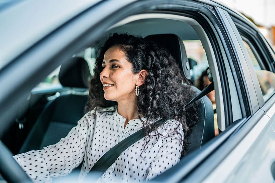 A woman with curly hair wearing a white polka dot shirt smiles while sitting in the driver’s seat of a car, holding the steering wheel and wearing a seatbelt—perhaps on her way to meet with one of Metro Detroit’s trusted independent car insurance agents. A woman with curly hair wearing a white polka dot shirt smiles while sitting in the driver’s seat of a car, holding the steering wheel and wearing a seatbelt—perhaps on her way to meet with one of Metro Detroit’s trusted independent car insurance agents.