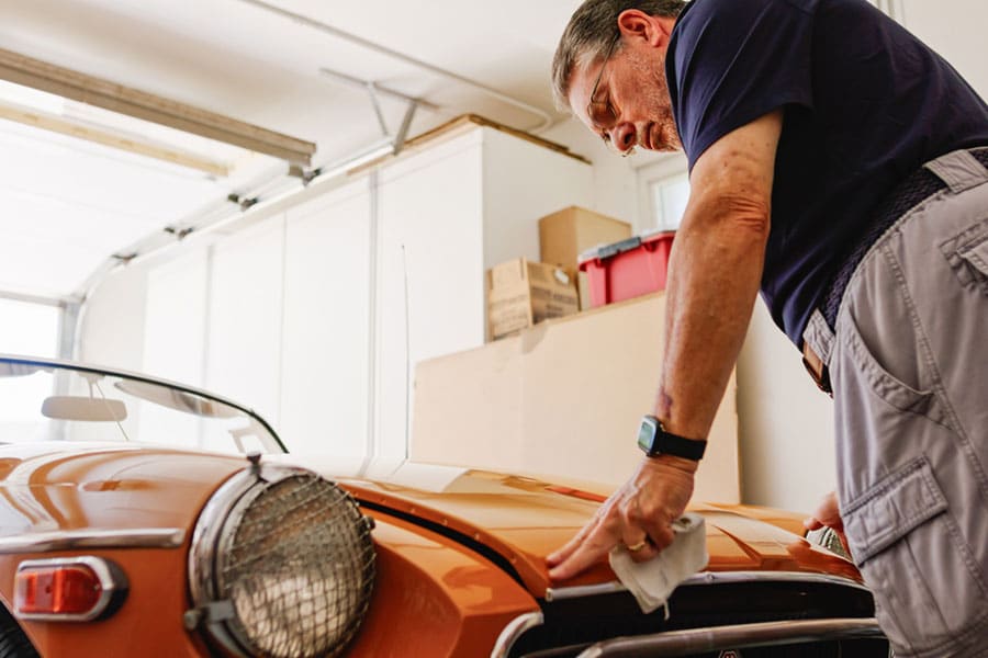 An older man wearing glasses polishes the hood of an orange classic car in his Metro Detroit garage, holding a white cloth and looking intently at his work.
