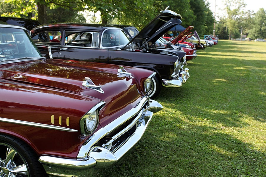 A row of classic cars with shiny chrome details is parked on grass at an outdoor car show in Metro Detroit, some with hoods open, trees and sunlight in the background—a perfect setting for enthusiasts to discuss Classic Car Insurance.