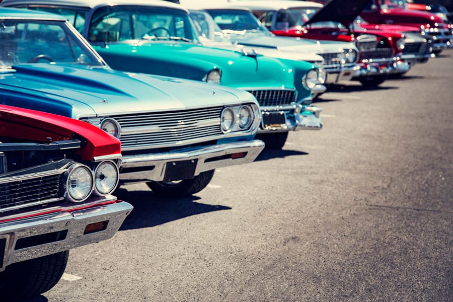 A row of colorful vintage cars is parked at an outdoor car show on a sunny day in Metro Detroit, with chrome details and classic designs visible in the front ends—a stunning showcase perfect for any classic car insurance enthusiast.