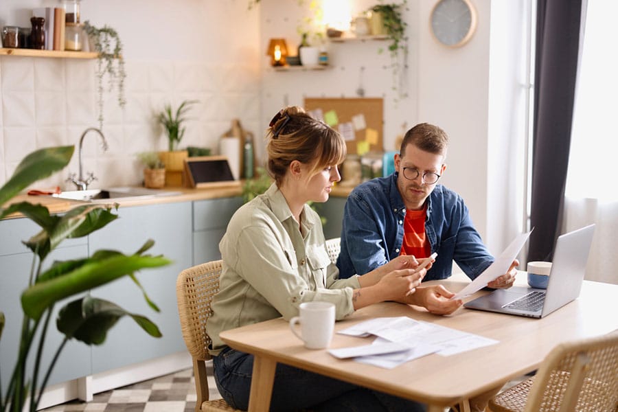 A man and woman sit at a kitchen table with paperwork, a laptop, and coffee mugs. They appear to be discussing something—perhaps reviewing options from independent home insurance agents in Metro Detroit—with the man holding papers and the woman gesturing. The kitchen is bright and modern. A man and woman sit at a kitchen table with paperwork, a laptop, and coffee mugs. They appear to be discussing something—perhaps reviewing options from independent home insurance agents in Metro Detroit—with the man holding papers and the woman gesturing. The kitchen is bright and modern.