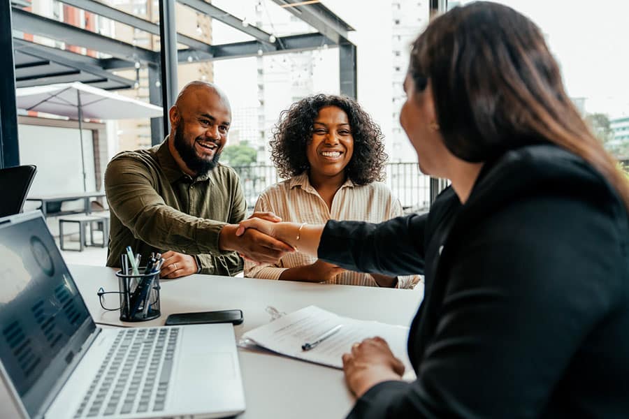 Three people sit at a table with a laptop and documents; two people smile as one shakes hands with a third person across the table during a meeting, reflecting the trusted partnership often found between clients and Independent Home Insurance Agents in Metro Detroit. Three people sit at a table with a laptop and documents; two people smile as one shakes hands with a third person across the table during a meeting, reflecting the trusted partnership often found between clients and Independent Home Insurance Agents in Metro Detroit.