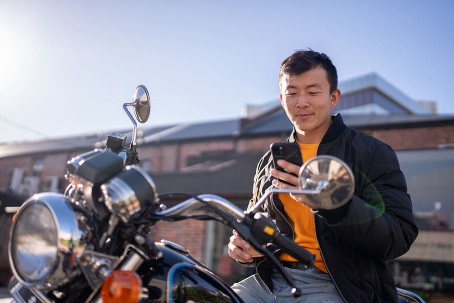 A person sitting on a motorcycle outdoors in Metro Detroit, wearing a black jacket and holding a smartphone, smiles while looking at the screen. Buildings and a clear sky are visible in the background, reflecting the freedom that comes with great motorcycle insurance options from independent agents.