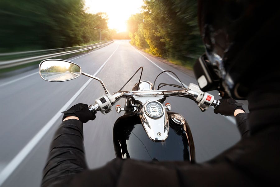 A person wearing a helmet rides a motorcycle down an empty road at sunset, with trees and guardrails blurred in motion on either side—a perfect scene for Metro Detroit independent agents to highlight the importance of reliable motorcycle insurance.